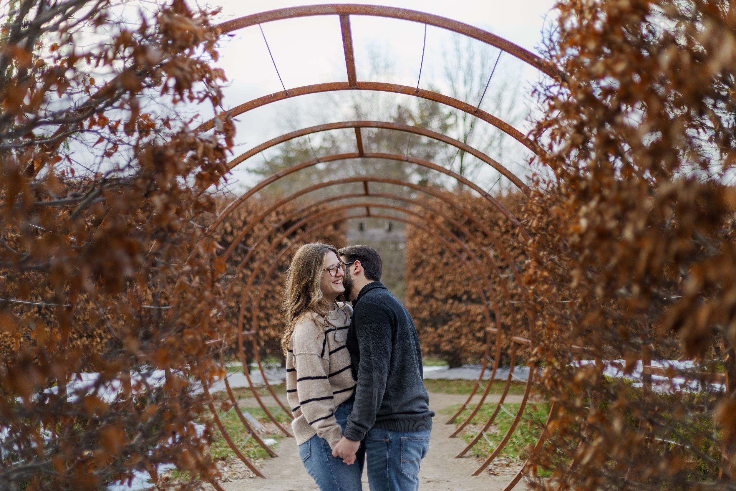 A couple embraces under an arched walkway surrounded by autumn foliage during a romantic photoshoot.
