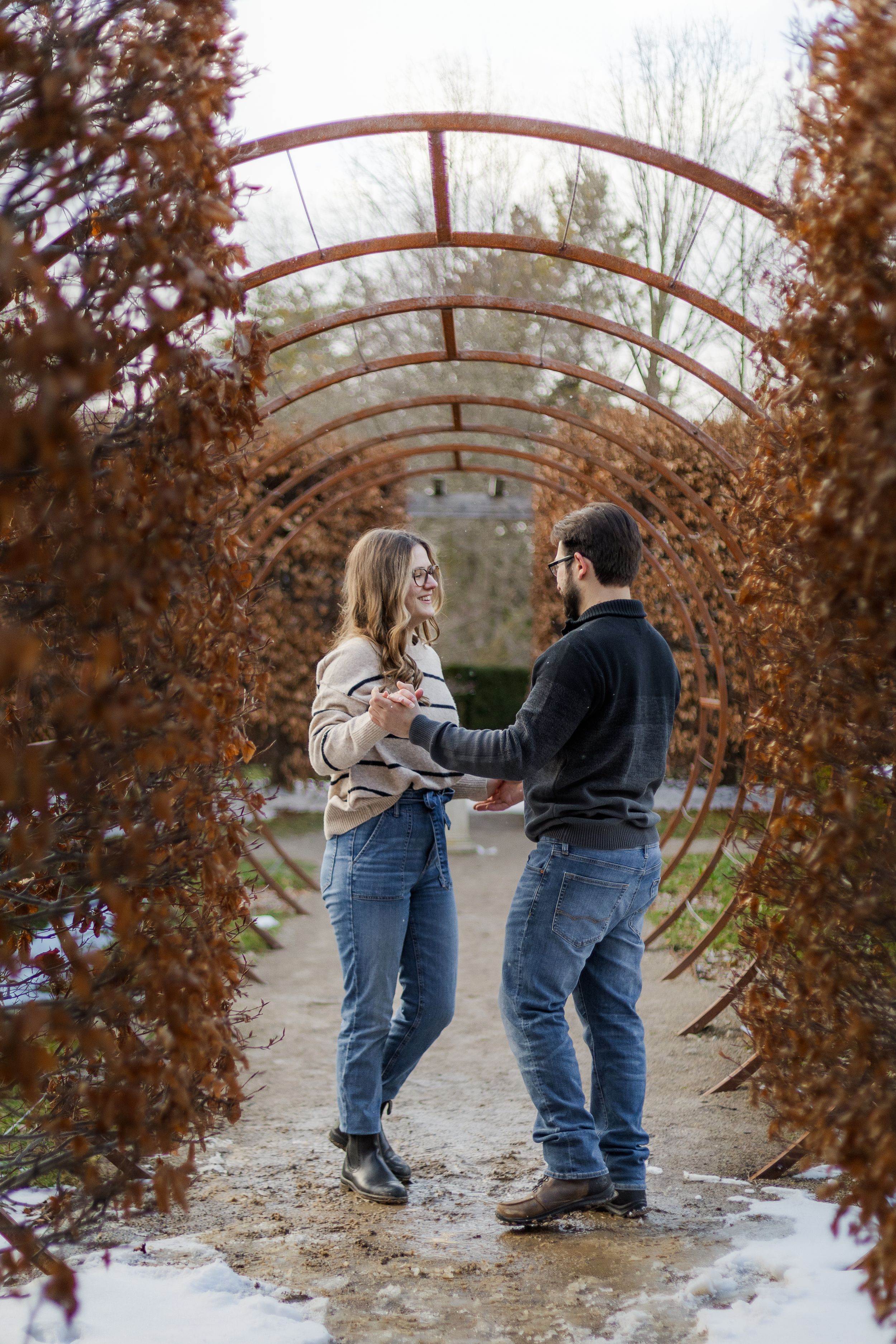 Two people in winter coats and jeans dance playfully under a curved metal archway surrounded by autumn foliage and snow.
