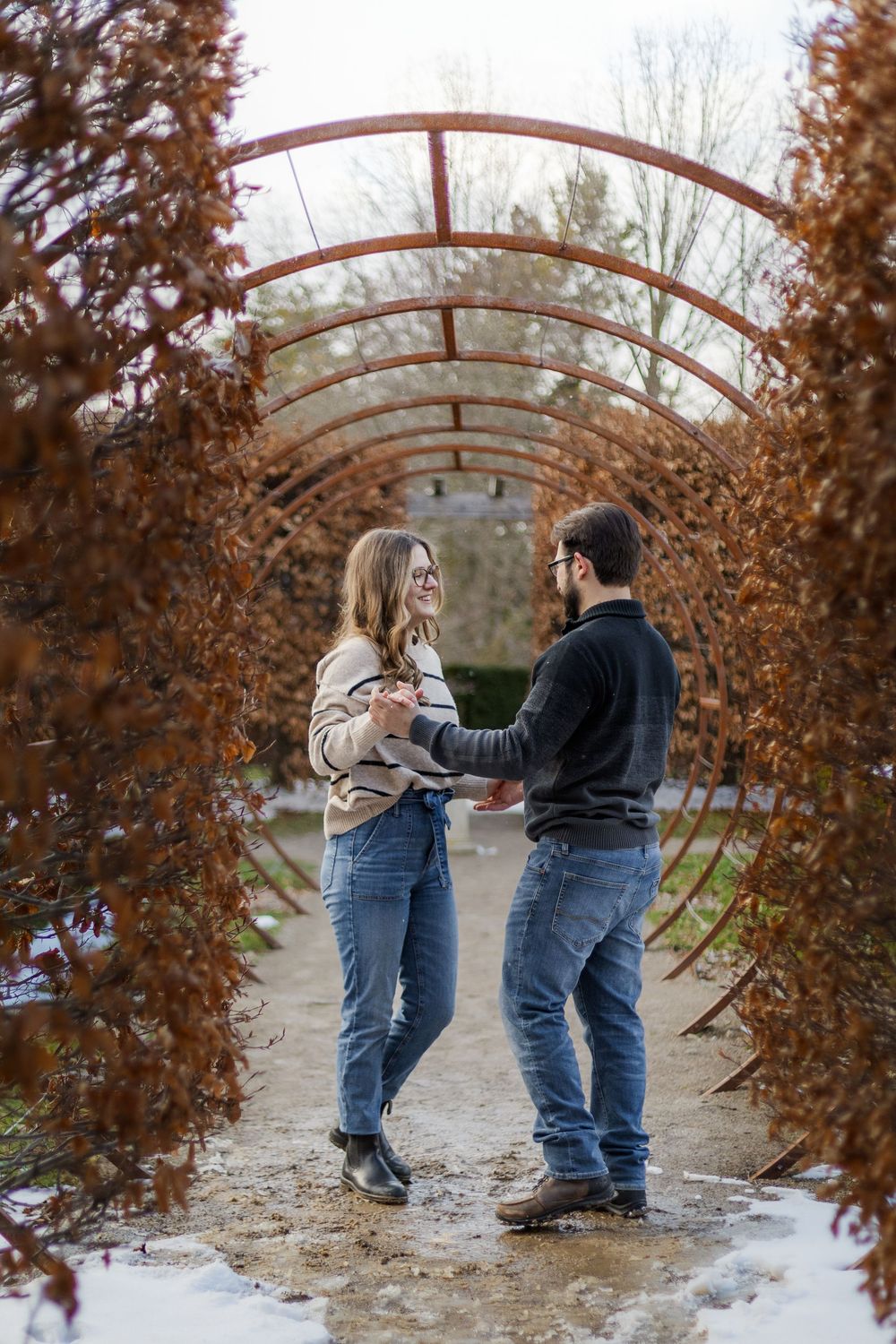 Two people in winter coats and jeans dance playfully under a curved metal archway surrounded by autumn foliage and snow.