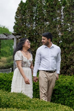 A couple shares a romantic moment while walking through a manicured garden with evergreen trees in the background.