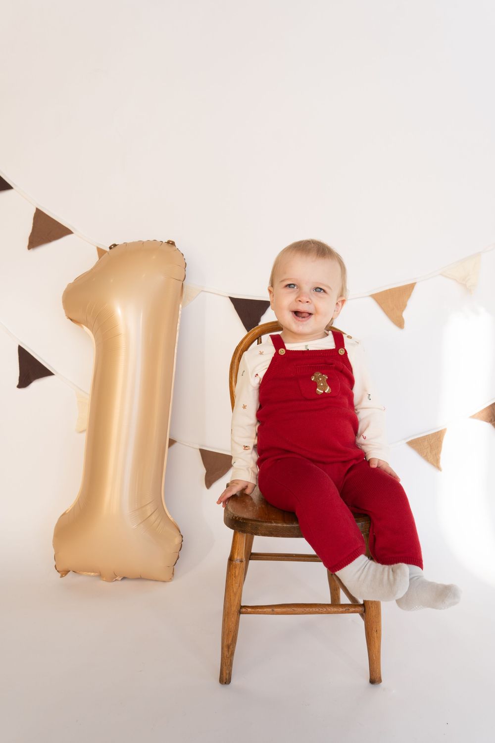 Baby in red overalls sits on wooden chair next to gold number one balloon.