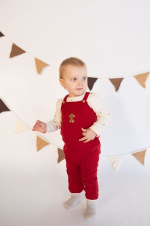 Happy baby in red corduroy overalls standing against white backdrop with party decorations.