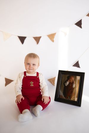 Baby in red overalls sits against white wall with decorative triangle banner bunting.