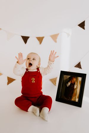 Baby in red overalls raises hands playfully against white backdrop with triangle banners.
