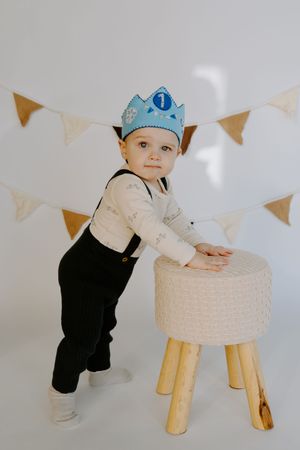 Small child in blue crown leaning against white ottoman with party bunting overhead.