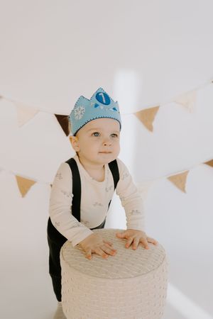 Baby wearing blue crown and black overalls leans on round white ottoman.