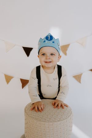 Smiling baby in blue crown and overalls poses with white ottoman under party banners.