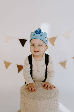 Happy baby in crown and overalls leaning on textured white ottoman under party flags.