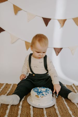 Small child in black overalls sits on striped rug with decorated white cake and festive bunting overhead.