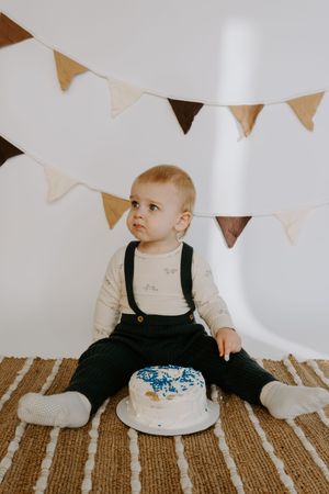 Baby in black overalls looks up at bunting while sitting next to white frosted cake on striped rug.