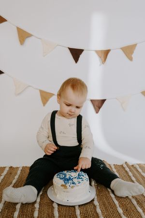 Baby explores white frosted cake with blue decorations while sitting on textured brown carpet.