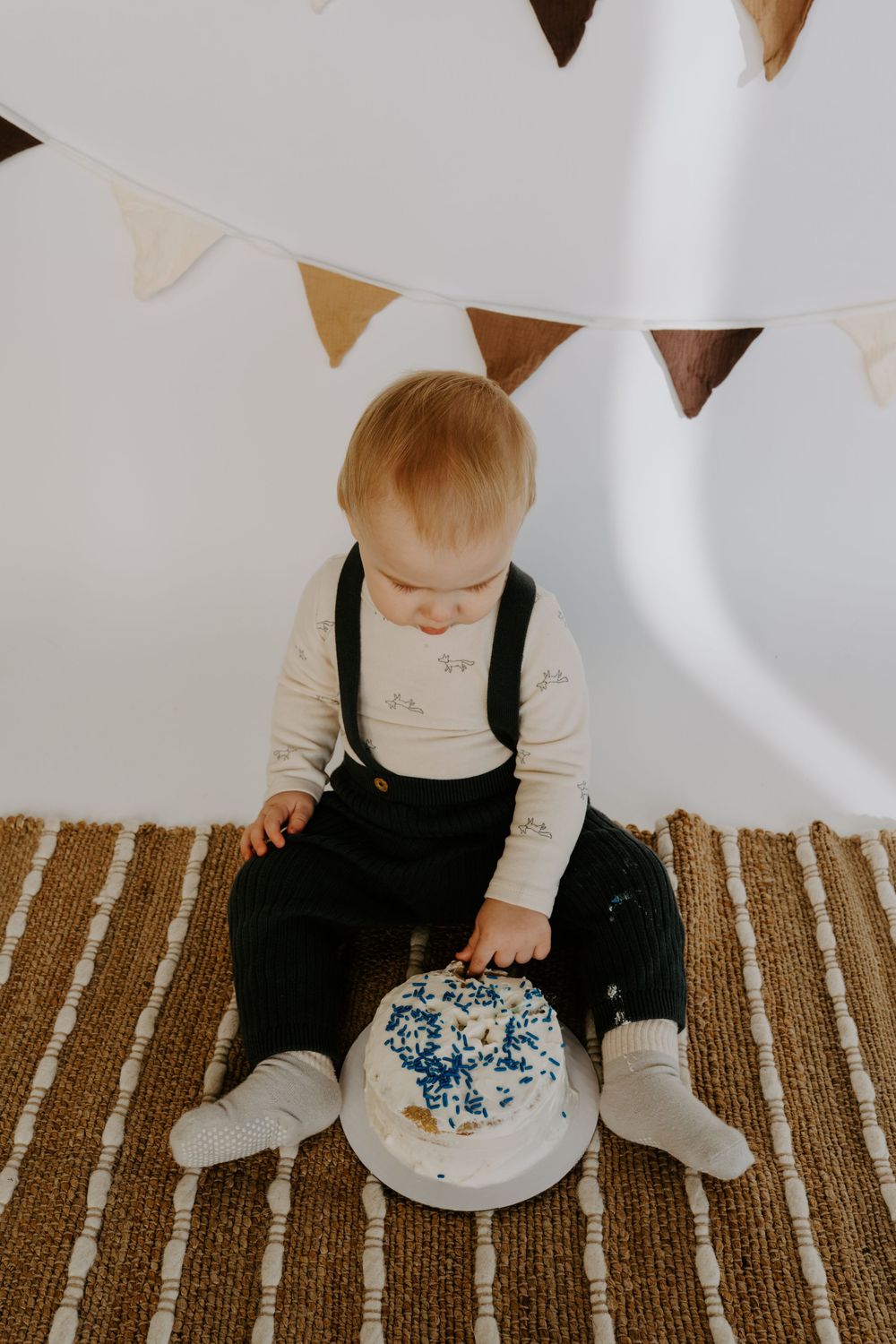 Small child in dark overalls examines white cake with blue sprinkles on striped floor.