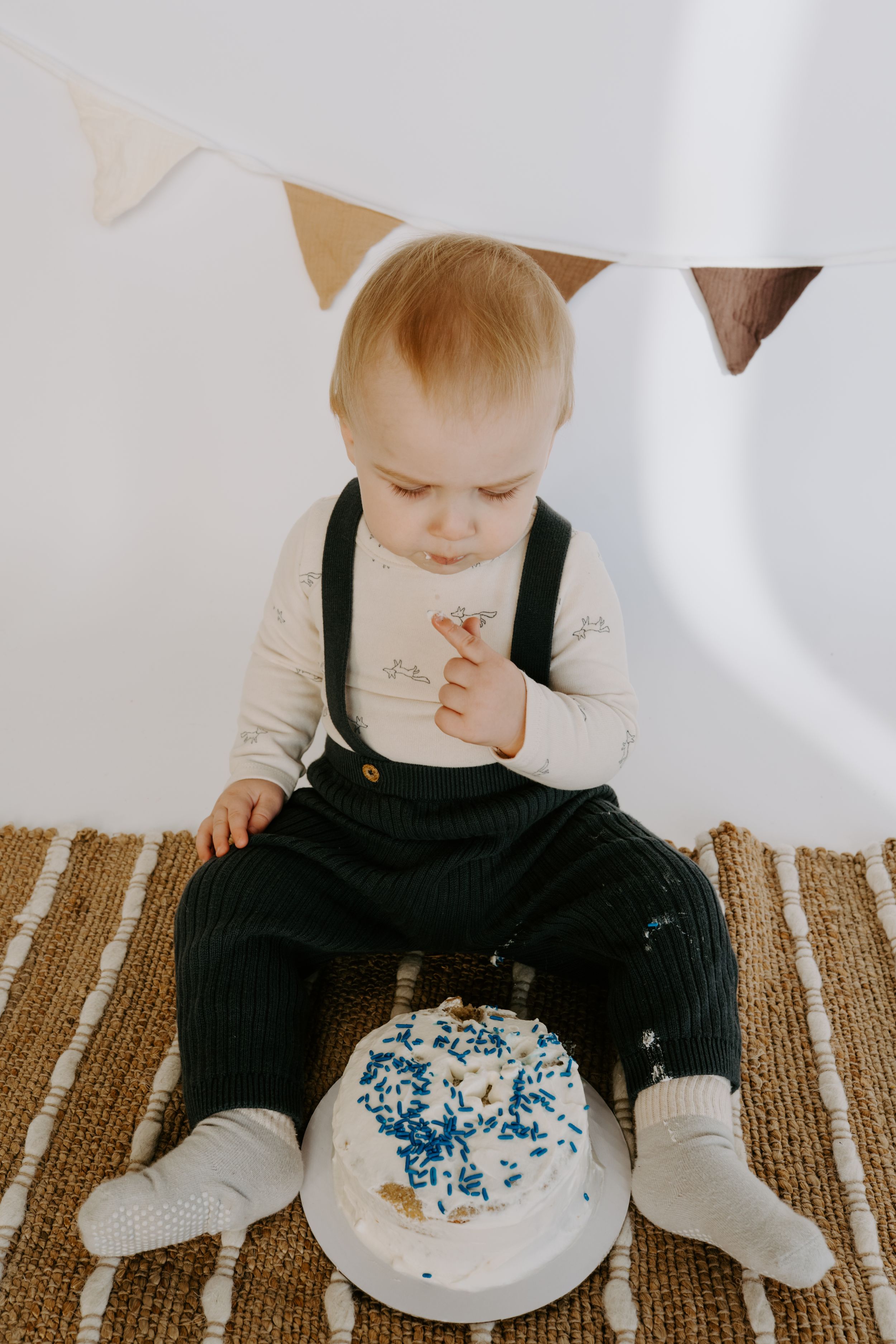 Baby in black overalls investigates white frosted cake during photo session on brown rug.