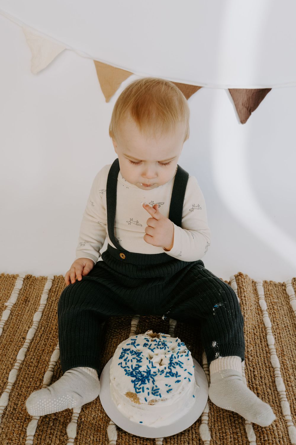 Baby in black overalls investigates white frosted cake during photo session on brown rug.