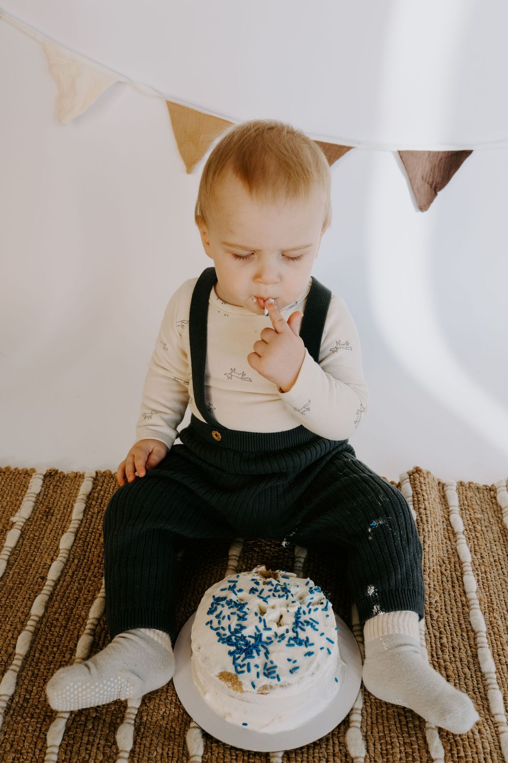 Baby in dark overalls reaches toward white cake with blue decorations under festive bunting.