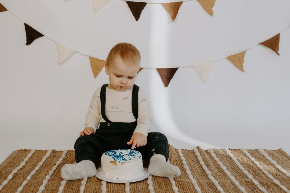 Small child in overalls and white shirt sits near decorated cake under hanging pennant banners.