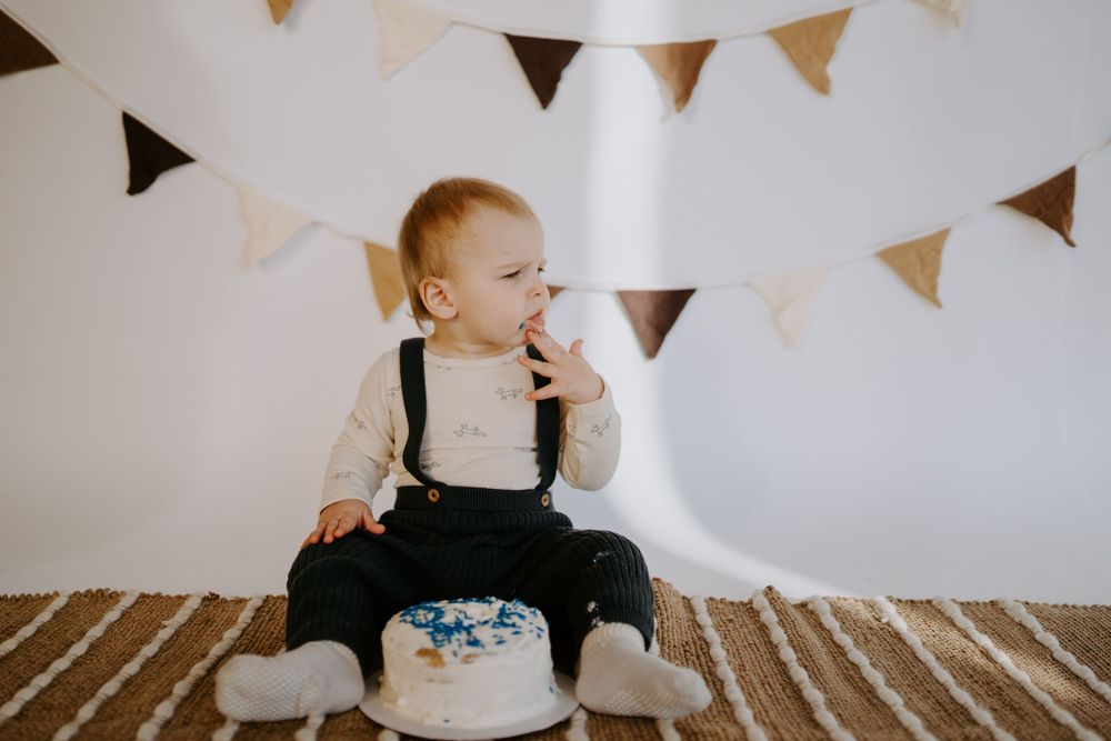 Small child in black overalls sits on brown carpet with white cake and decorative banners.