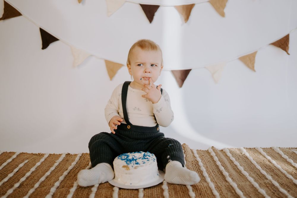 Baby in dark overalls explores white frosted cake during celebration photoshoot with hanging bunting.