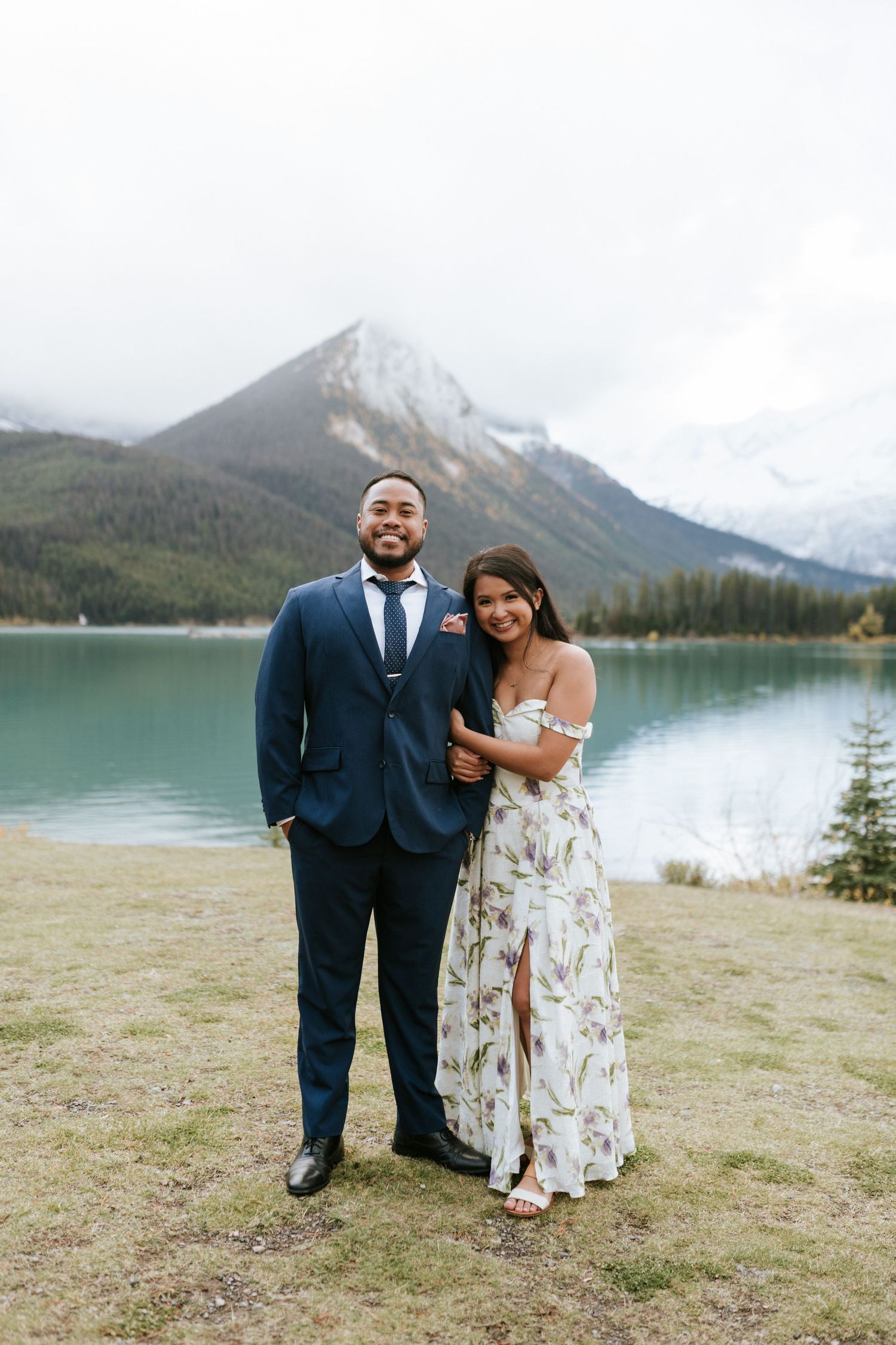 A couple poses together in formal attire against a scenic mountain lake backdrop in a romantic outdoor setting.