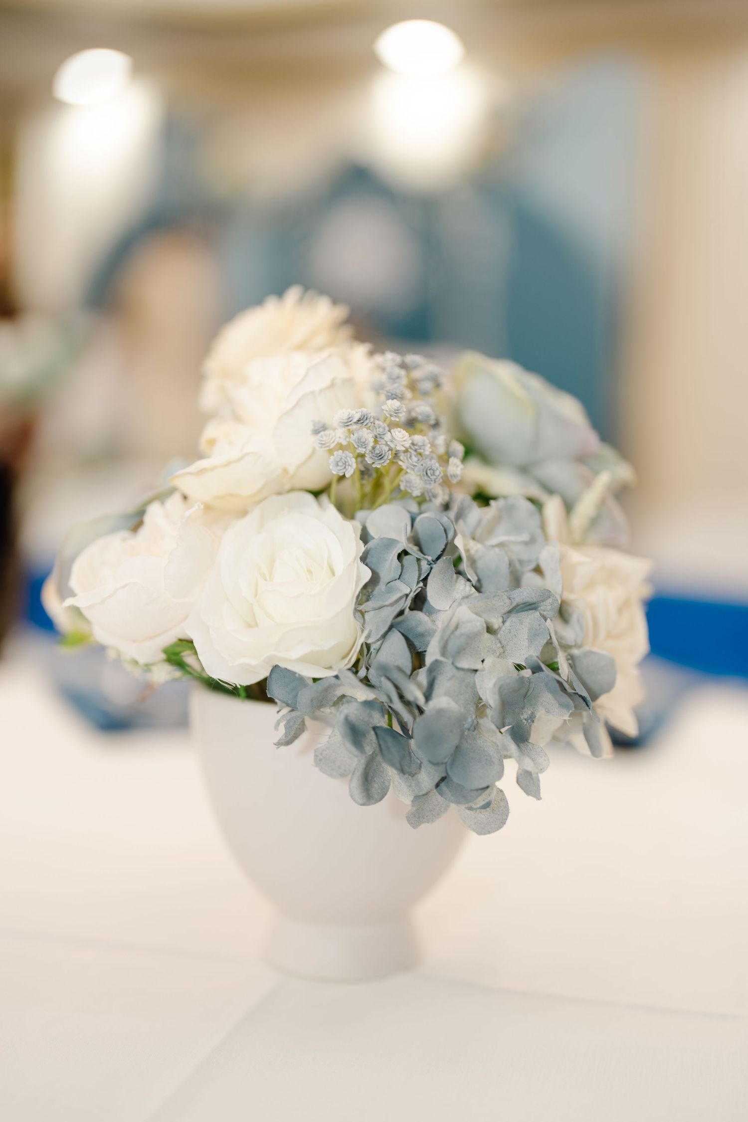White vase with white roses and blue hydrangeas centerpiece on a table.