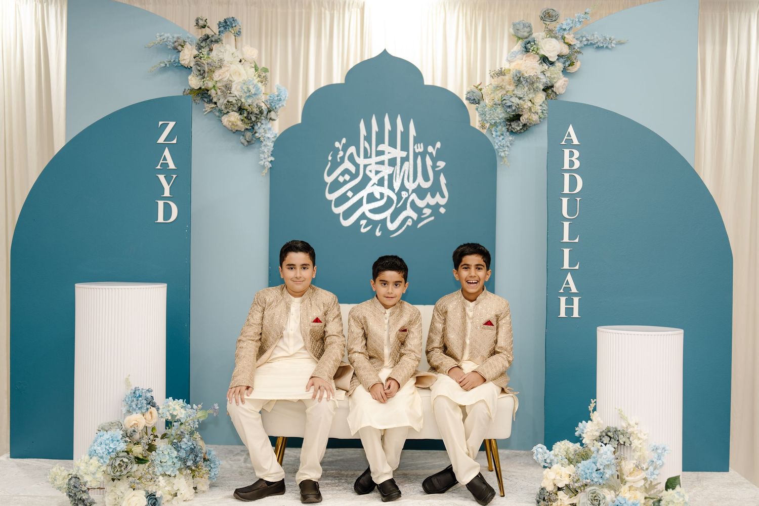 Three boys in gold sherwanis posed at a blue Islamic-themed celebration backdrop.