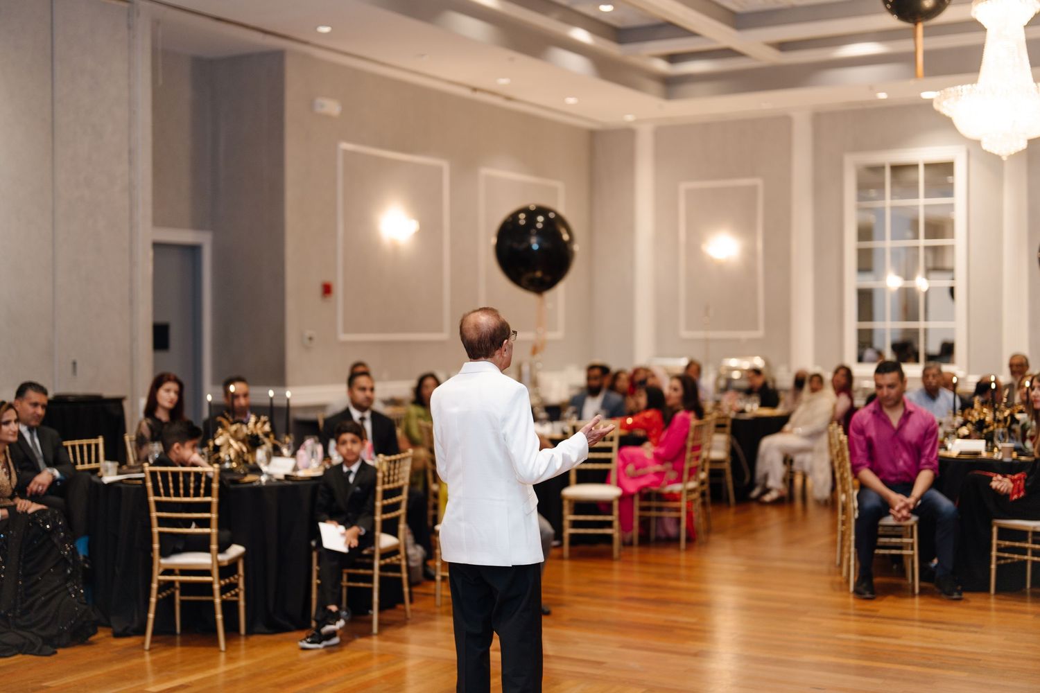 Man in white jacket addresses guests at an elegant black and gold themed birthday celebration in a ballroom.