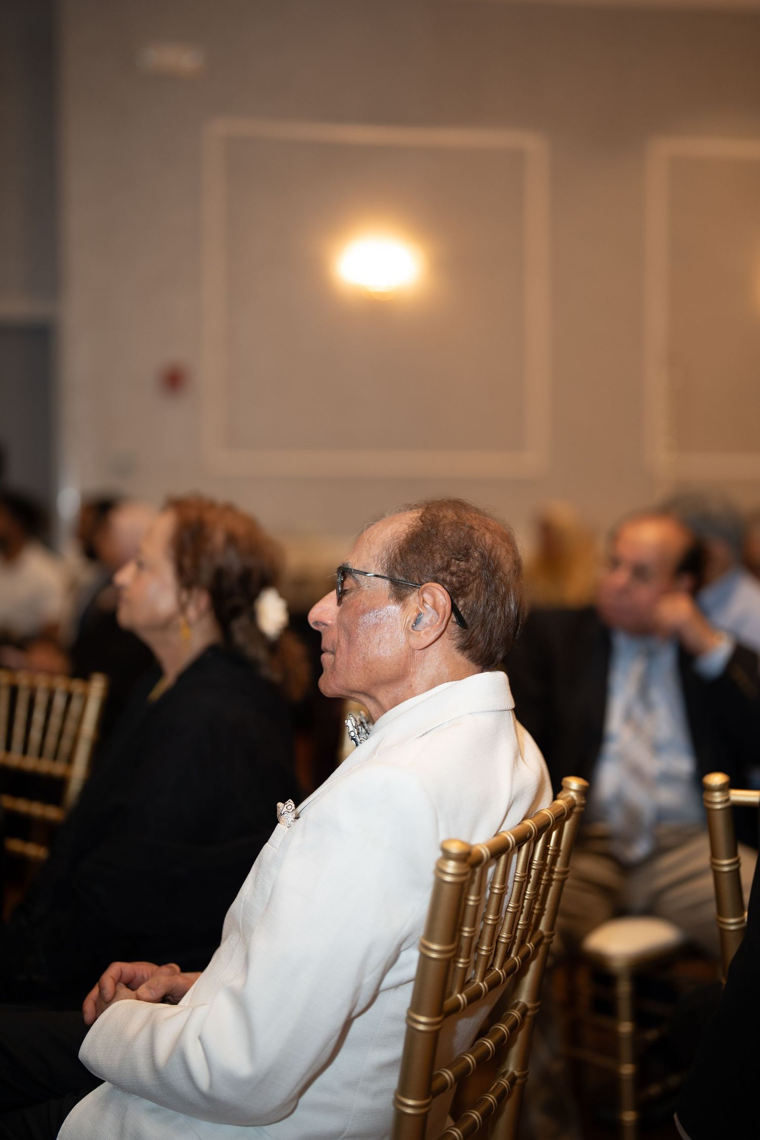 Elderly man in white jacket seated at an indoor celebration event.