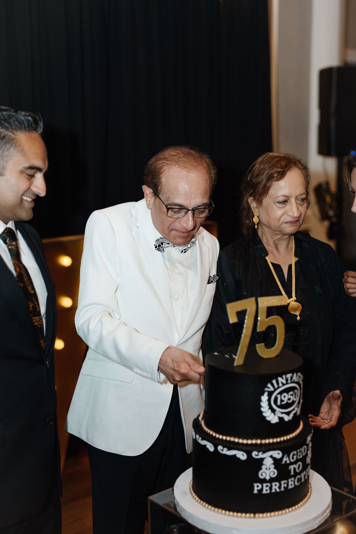 Elderly man in white tuxedo cuts a black and gold 75th birthday cake with woman beside him.