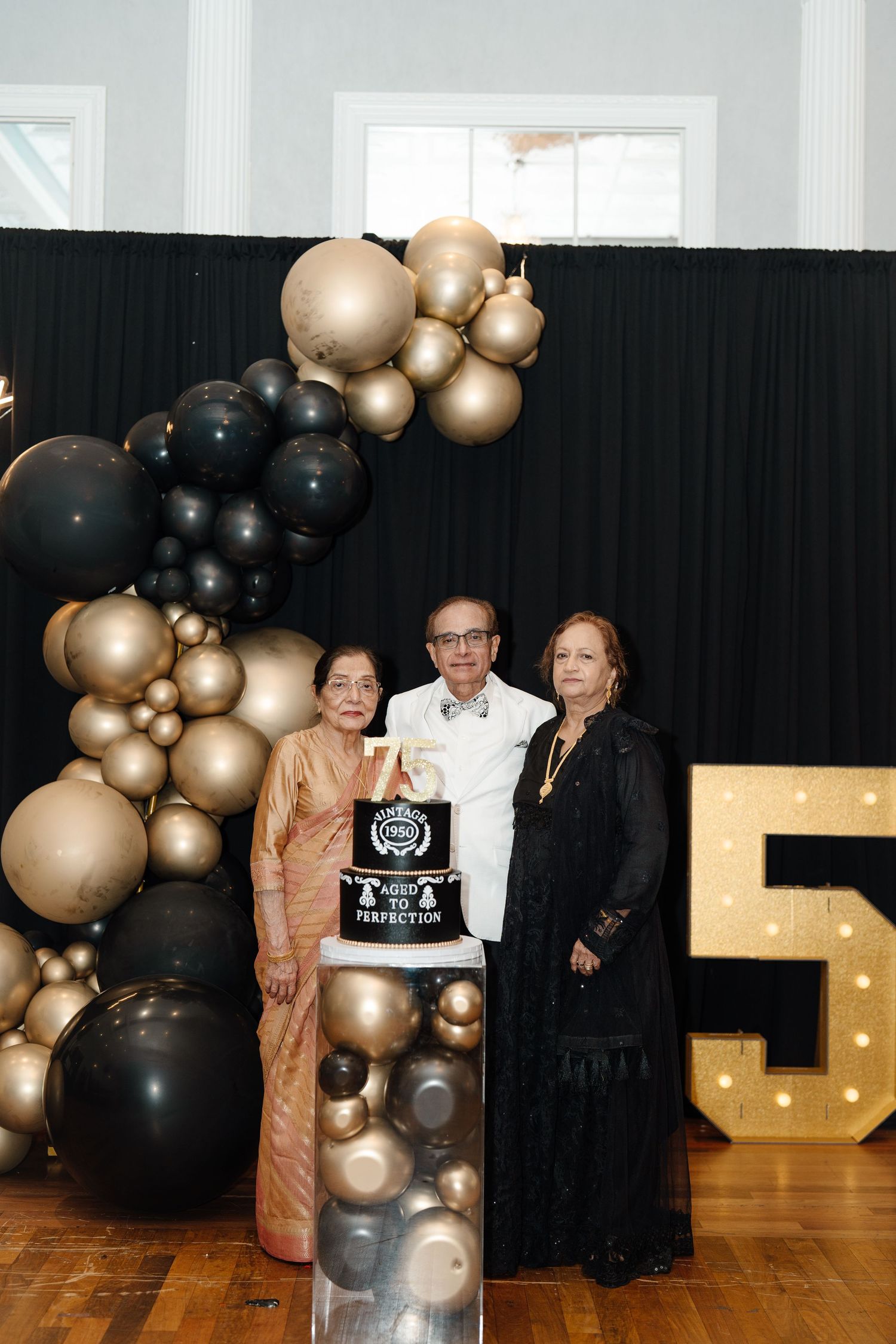 Three people pose with a black and gold 75th birthday cake in front of balloon arch decor.