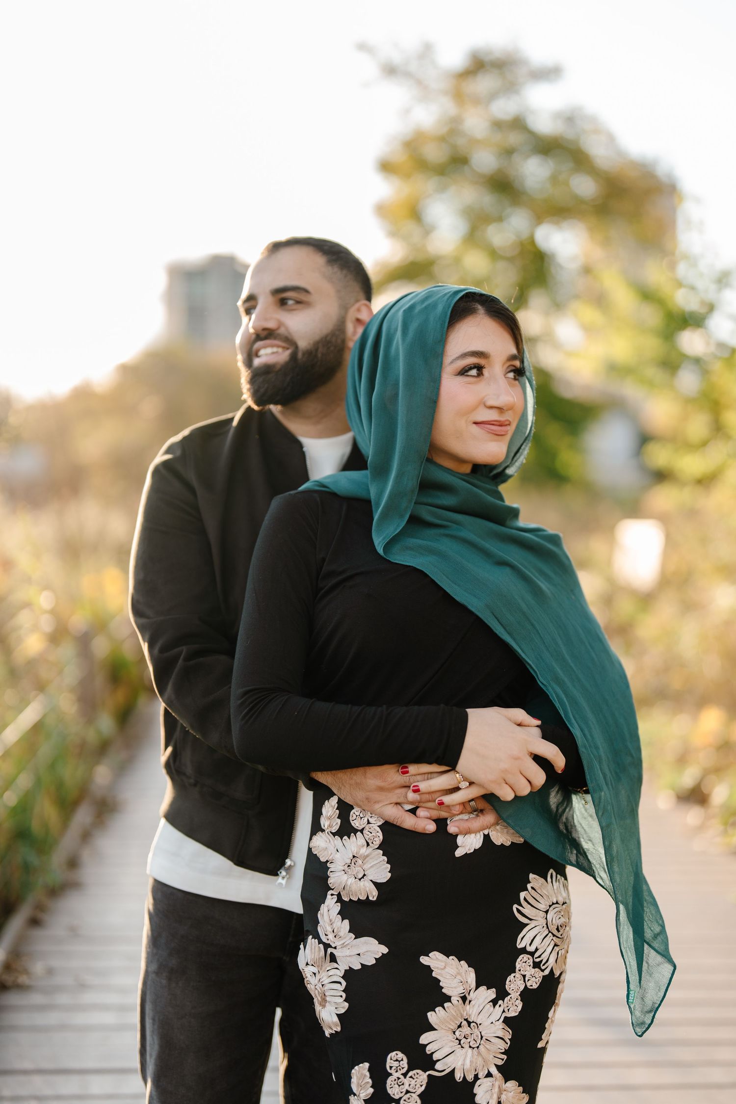 Couple embracing on boardwalk at golden hour, woman wearing teal hijab and floral skirt.
