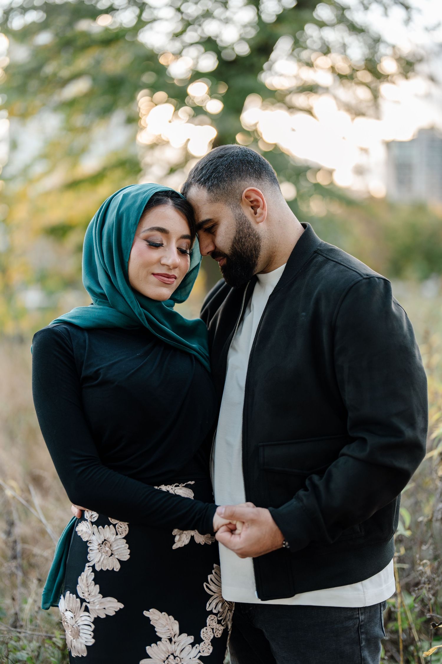 A couple in a tender embrace outdoors, woman wearing a green hijab and floral skirt, man in a black jacket.