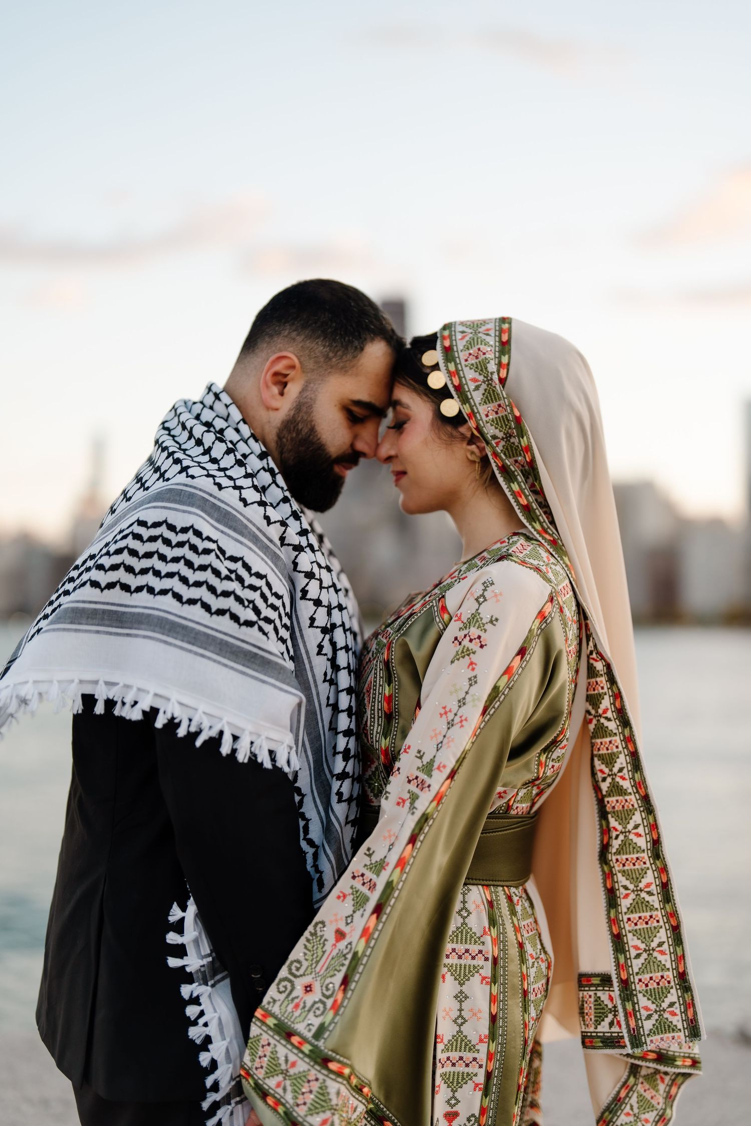 A couple in traditional Palestinian attire touching foreheads romantically with Chicago skyline in background.
