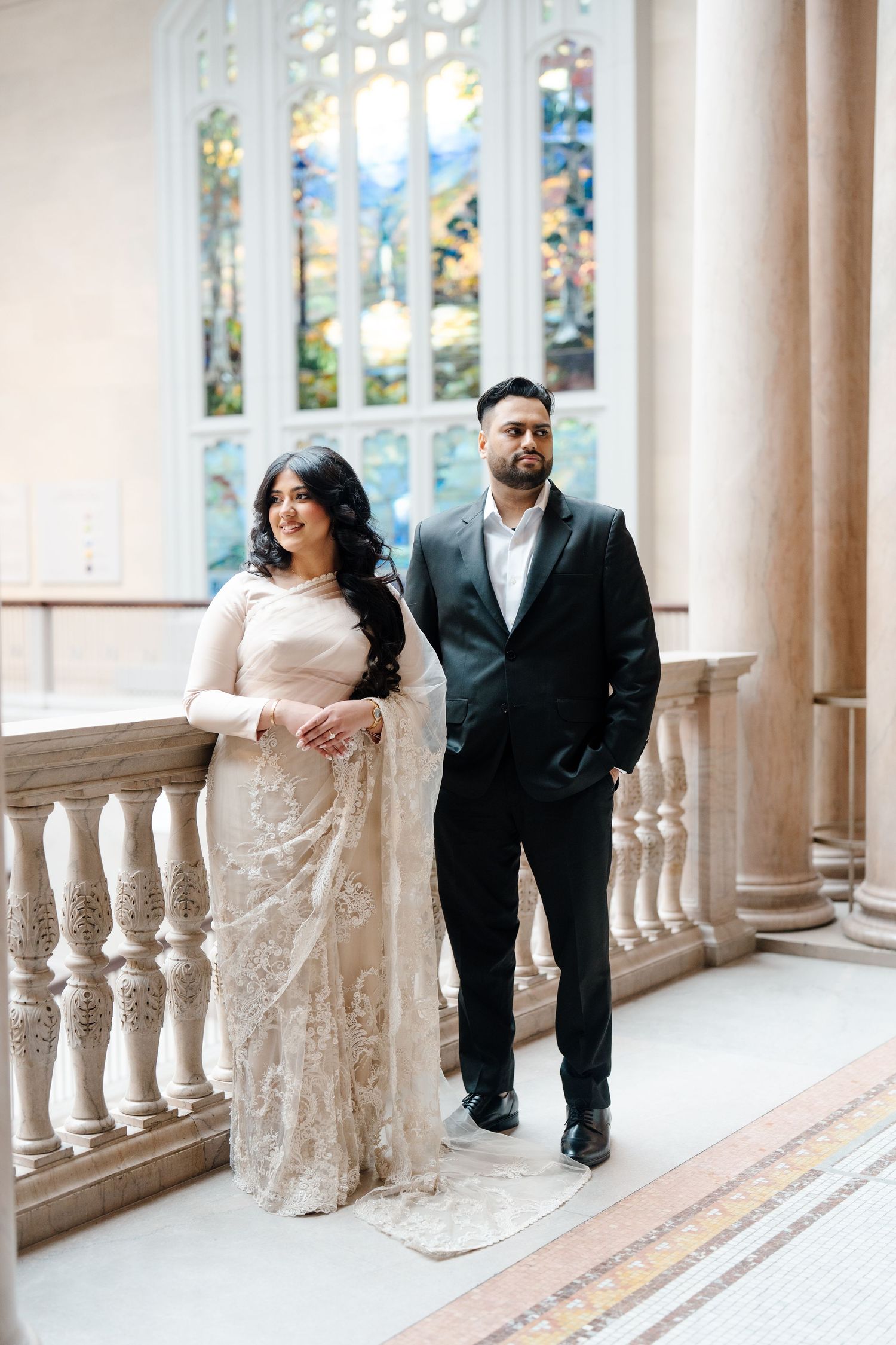 Couple standing by ornate balustrade with stained glass window, woman in white lace saree, man in black suit.