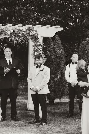 Groom in beige suit and bow tie waits at floral wedding arch during outdoor ceremony.