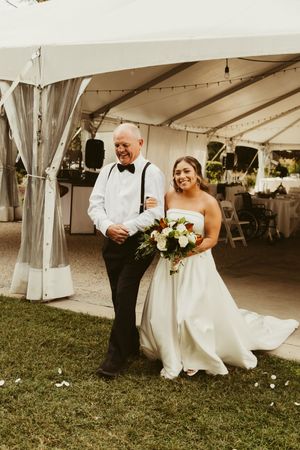 Bride in white gown walks with father under tent entrance holding bouquet at wedding.