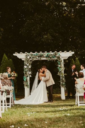 Bride and groom share first kiss under floral arch at outdoor garden wedding ceremony.
