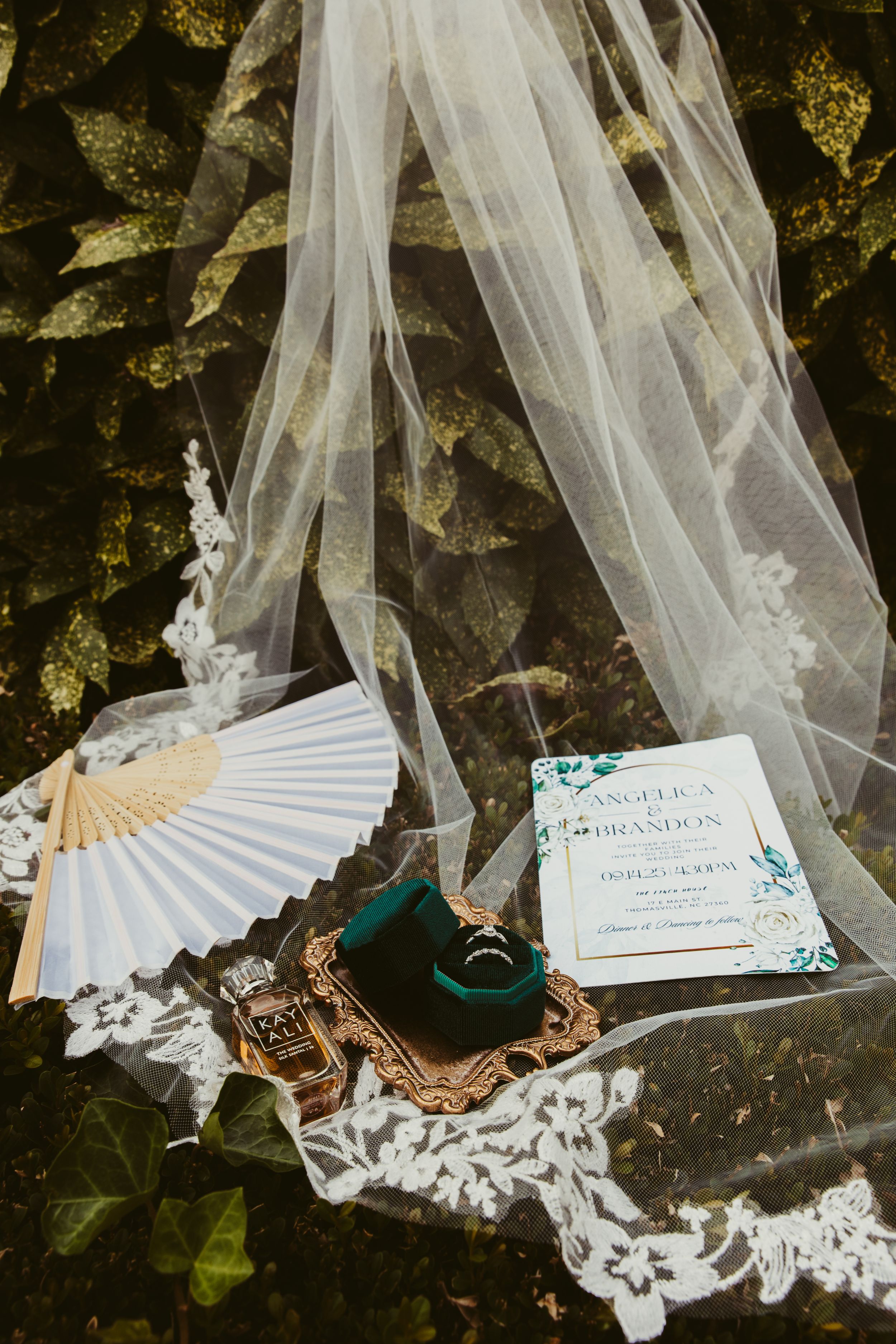Wedding flat lay with veil, invitation, emerald ring box, and perfume on lace.