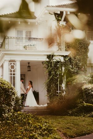 Bride and groom hold hands on porch of white ivy-covered estate wedding venue.