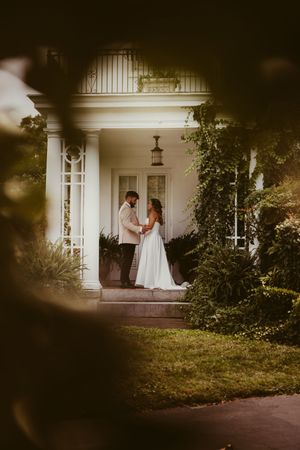 Bride and groom face each other on steps of elegant white columned mansion.