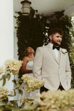 Bride laughs behind groom in tan tuxedo on ivy-covered porch with hydrangeas.