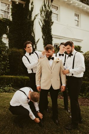 Groomsmen in black suspenders and bow ties help groom in tan suit prepare outside ivy-covered venue.