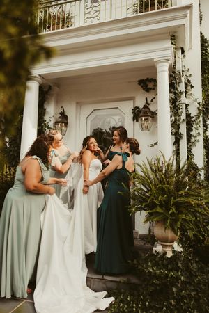 Bridesmaids in sage and emerald green dresses help bride with veil on steps of white colonial venue.