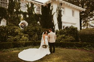 Bride in long cathedral train gown and groom share intimate moment in garden of ivy-covered estate.