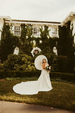Bride in elegant white gown with cathedral train poses with parasol and bouquet before ivy-covered mansion.