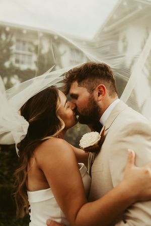 Bride and groom share a kiss under flowing wedding veil in a romantic close-up portrait.
