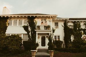 Bride and groom stand on balcony of grand ivy-covered white colonial wedding venue at golden hour at The Finch House in Thomasville, NC.