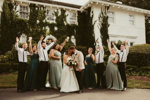 Wedding party in sage green dresses and black suspenders celebrate with raised hands outside white mansion.