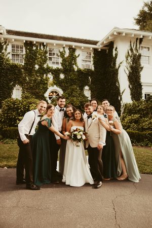 Bride and groom pose with wedding party in green gowns and suspenders outside ivy-covered white estate.