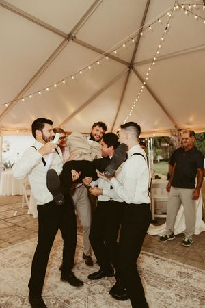 Groomsmen in white shirts playfully lift groom on dance floor at wedding reception.