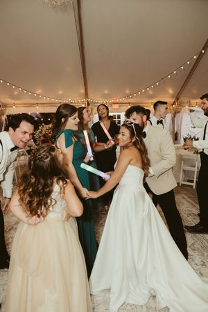 Bride dances with bridesmaids holding glow sticks at lively tented wedding reception.